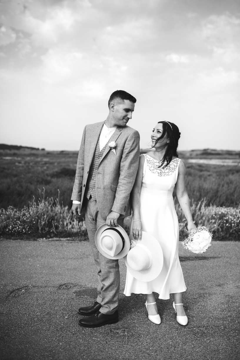 photo noir et blanc, le couple est dos à dos, où la mariée se retourne pour regarder son mari, elle tient le bouquet dans ses mains avec dans l'autre un chapeau, et lui, tient un chapeau